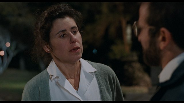 A distressed woman in a white shirt and cardigan talks to a man in the evening. The background appears blurred with lights.