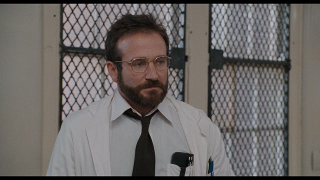 A man with a beard wearing glasses and a white lab coat stands in a room with metal mesh windows.
