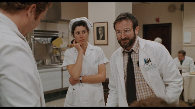 A scene from the movie featuring three characters in a hospital setting, with one man wearing glasses and a white coat talking to a nurse and another staff member.