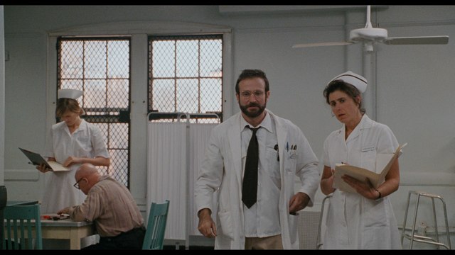 A scene from a hospital setting featuring a doctor in a white coat and glasses, accompanied by a nurse in a white uniform. In the background, another nurse is attending to paperwork, while a patient is seated at a table.