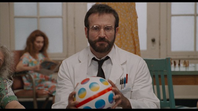 A man in a doctor's coat holds a colorful ball while sitting in a room with patients. He has a beard and glasses and appears focused. In the background, another patient sits holding a magazine.