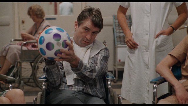 A scene from a movie featuring a man in a wheelchair, holding a colorful ball, with a nurse beside him and other patients in the background.