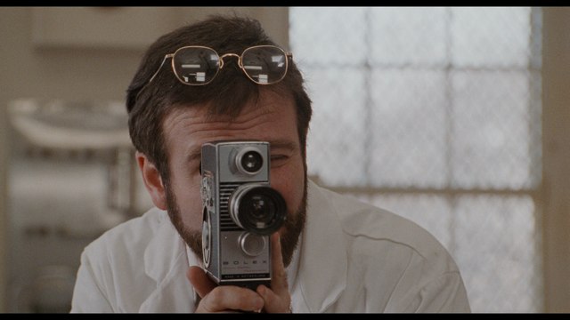 A man with glasses holds a vintage video camera, aiming it toward the viewer. The setting features a window with a patterned design in the background.