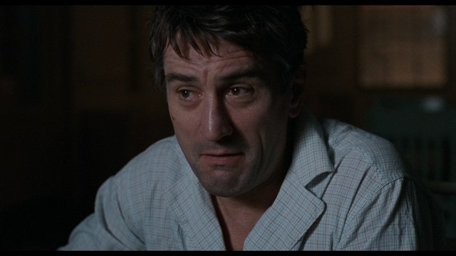 A close-up image of a man looking concerned or uneasy, wearing a light checkered shirt. The background appears dimly lit, suggesting an indoor setting.