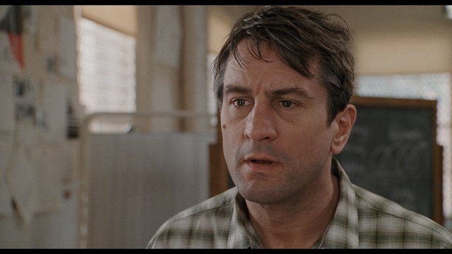 Close-up of a man with short hair and a concerned expression, wearing a plaid shirt, in a setting resembling a medical facility.