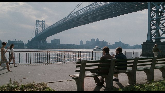 Two men sitting on a park bench by a river, with the city skyline and a bridge in the background.