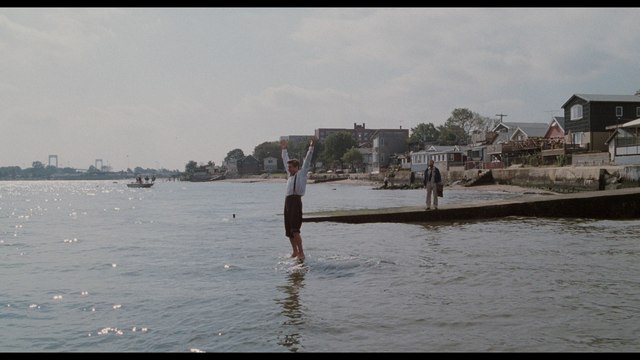 A man stands in shallow water with arms raised, while another person stands on a dock in the background, surrounded by houses by the shore.