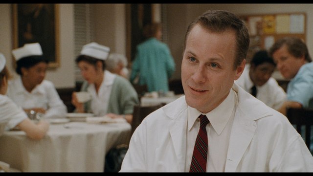 A smiling doctor in a white coat sits at a table in a hospital cafeteria, while nurses and staff in the background engage in conversation.