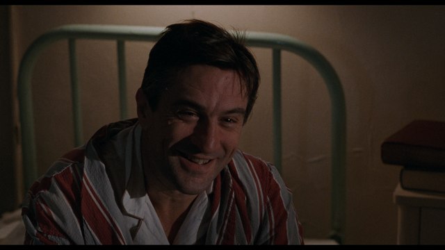 A scene from a film showing a man in a striped pajama shirt smiling warmly in a dimly lit room with a green metal bed frame in the background.