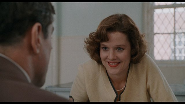 A woman with brown hair styled in soft waves smiles while sitting at a table, engaging in conversation with a man off-screen.