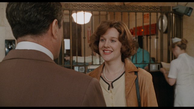 A woman with curly hair smiles while speaking to a man in a brown suit at a reception area in a hospital-like setting.