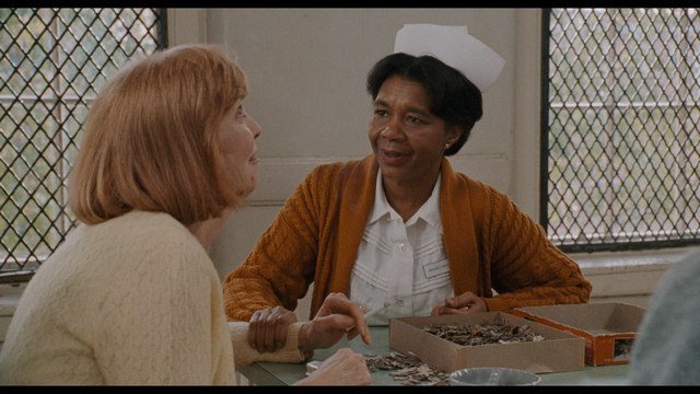 A scene from a film showing two women engaged in conversation at a table, with one woman wearing a nurse's uniform.