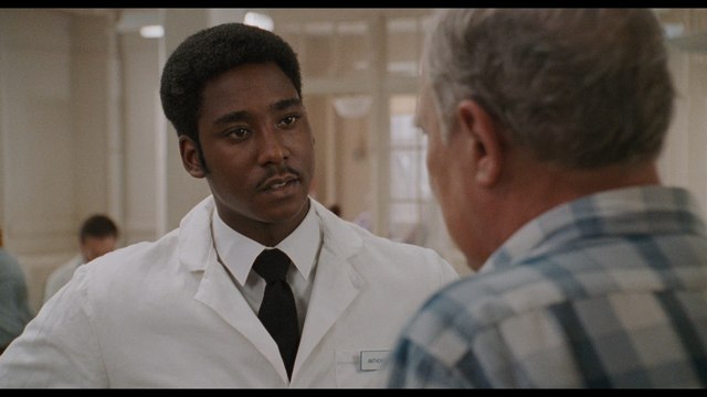 A scene from a hospital where a character in a white coat is talking to an older man wearing a patterned shirt.