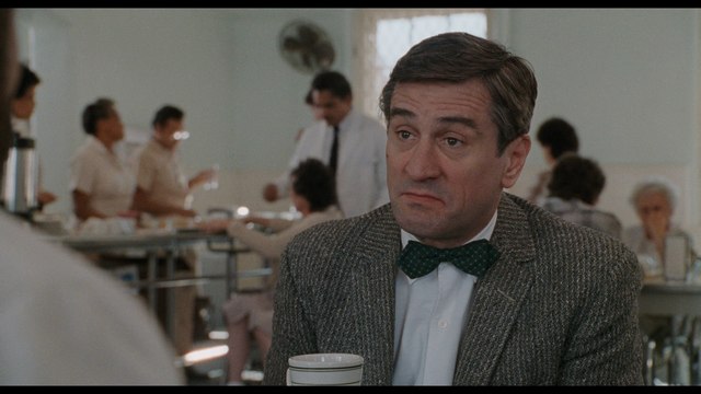 A scene from a restaurant featuring a male character wearing a bow tie and textured jacket, sitting at a table with a cup in hand while appearing contemplative. Background includes other characters and a bustling atmosphere.