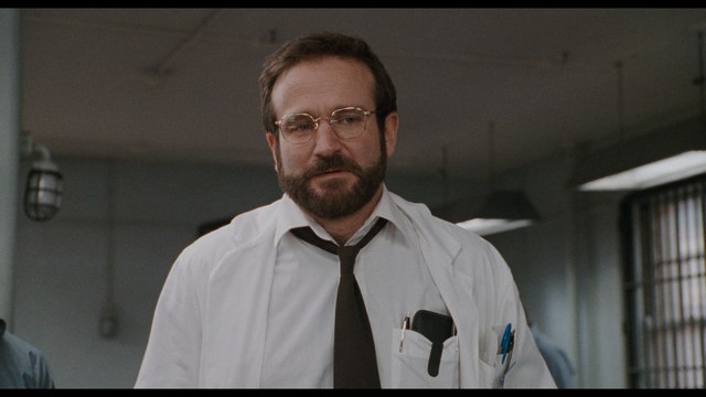 A scene from a movie featuring a man in a white lab coat and glasses, looking serious in a clinical setting.