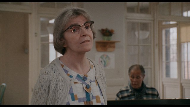 A middle-aged woman in a patterned blouse and cardigan looks directly at the camera, with glasses on her face and short gray hair. In the background, an older man sits at a piano.