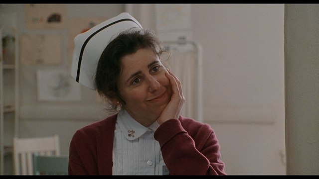A nurse with dark hair, wearing a white nurse's cap and a red cardigan, is smiling warmly while resting her chin on her hand in a softly lit room.