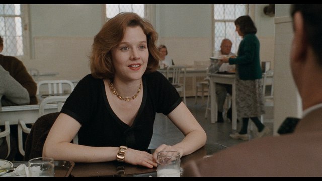 A woman with curly hair, wearing a black shirt and necklace, smiles while sitting at a table in a café surrounded by other patrons.