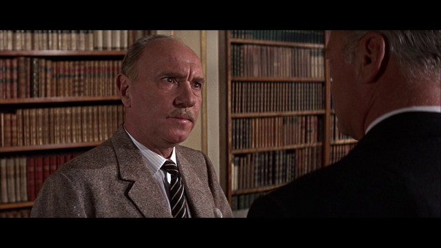 A man with large white wings stands in a room, wearing a dark suit, with a serious expression on his face. Bookshelves and curtains are visible in the background.