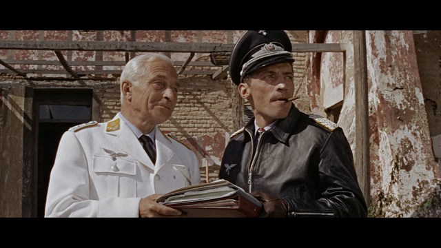 Two young men are looking at something off-camera, standing inside a building with bright lighting and seating areas visible in the background.