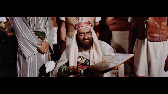 A close-up of an elderly man with a beard and white attire, appearing serious and contemplative, set against a colorful, futuristic background.