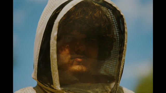 A serious-looking cardinal stands in front of a wooden backdrop with colorful posters related to Catholicism, holding a microphone at a press conference.