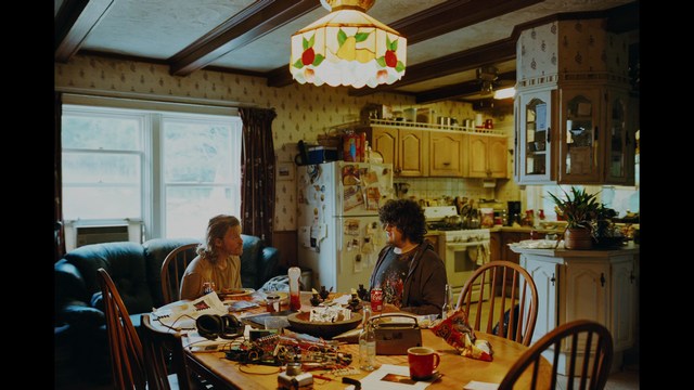 A woman with a pensive expression rests her chin on her hand, sitting at a table with kitchen appliances and items in the background.