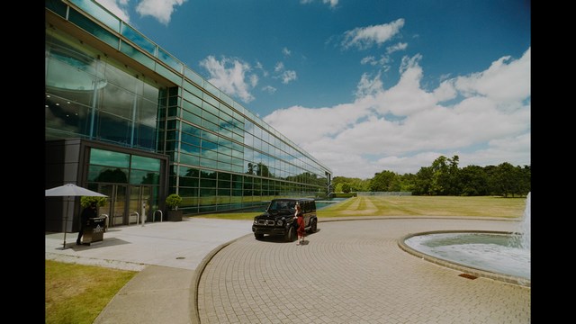 A man in a white suit and hat stands at a doorway, smiling, with a suburban landscape in the background.