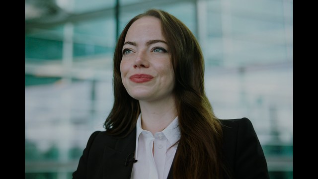 A man with large, feathered angel wings stands in a room, wearing a black suit and looking directly at the viewer.