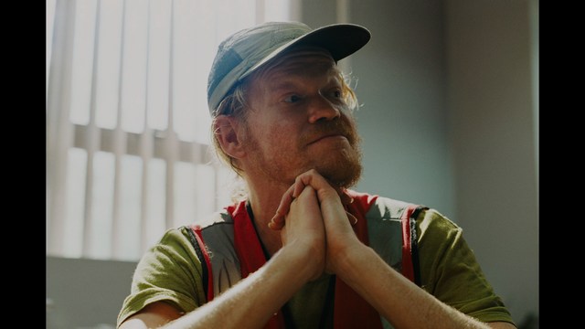 A scene featuring a man with short hair and a beard, wearing a brown jacket and purple shirt, standing outside a restaurant with bright yellow walls and a cartoon cow logo.