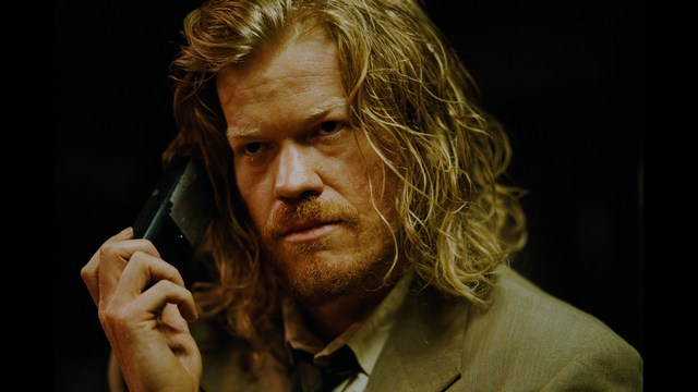 A golden trophy statue of a goblin-like character positioned on a wooden table in an office setting, with large windows and a building facade visible in the background.