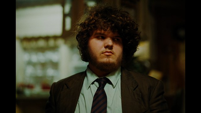 Two young men are looking at something off-camera, standing inside a building with bright lighting and seating areas visible in the background.