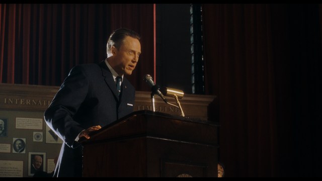 A man with a beard and mustache smiles warmly while standing on a boardwalk by the beach, with colorful flags overhead and a large building in the background.