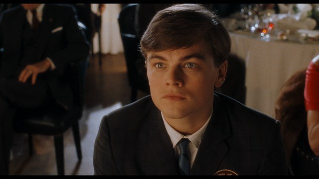 A serious-looking cardinal stands in front of a wooden backdrop with colorful posters related to Catholicism, holding a microphone at a press conference.