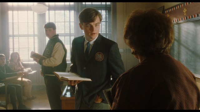 A woman with a pensive expression rests her chin on her hand, sitting at a table with kitchen appliances and items in the background.