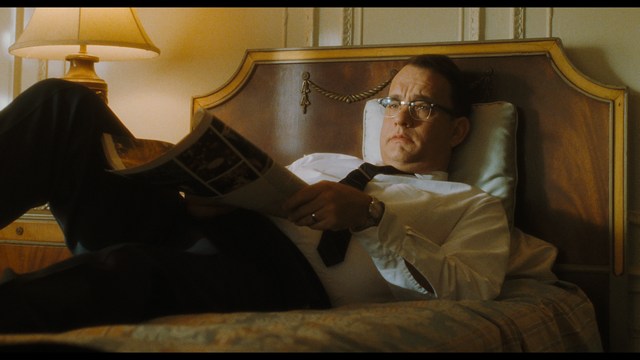 A man with large, feathered angel wings stands in a room, wearing a black suit and looking directly at the viewer.