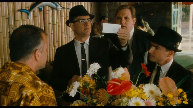 A golden trophy statue of a goblin-like character positioned on a wooden table in an office setting, with large windows and a building facade visible in the background.