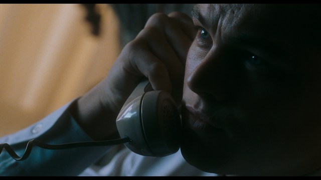 Close-up of a man with a serious expression, seated in a dimly lit area, with soft lighting from background lamps.