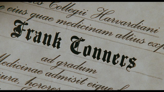 Two characters sitting at a table, one holding a small statue, with a surprised expression. The background features vibrant colors and patterns.