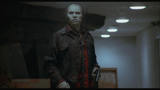 A menacing figure wearing a dark jacket stands in a dimly lit hallway, holding a gun and a chain.