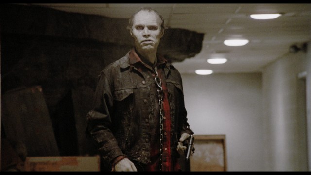 A sinister figure stands in a dimly lit hallway, wearing a tattered jacket, a blood-stained shirt, and holding a gun. The person has pale skin and appears menacing.