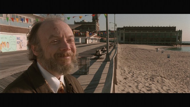 A smiling man with a beard stands on a wooden boardwalk near the beach, with colorful flags in the background and a building visible in the distance.