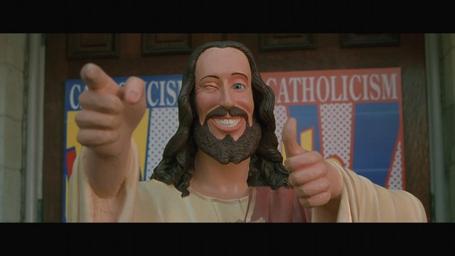 A large statue of a smiling man with long hair and a beard, giving a thumbs-up gesture, set against a background featuring colorful religious-themed posters.