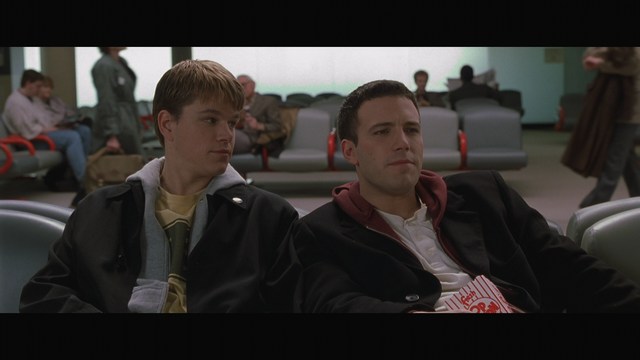 Two male characters sitting in a waiting area, one wearing a black jacket and the other in a maroon hoodie, with popcorn in hand.