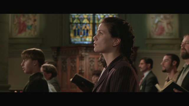 Scene from a church where a group of people, including a woman in a blazer and a young boy, are attending a service, facing forward with a stained glass window in the background.