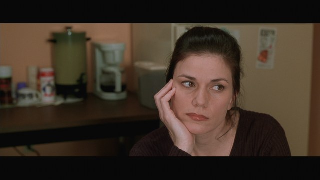 A woman with long dark hair resting her chin on her hand, looking pensively off to the side. In the background, various kitchen appliances and items are visible.