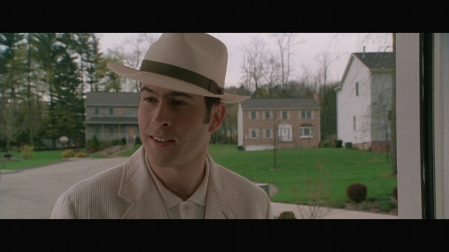 A man in a white suit and hat stands at the entrance of a house, smiling, with a suburban neighborhood visible in the background.