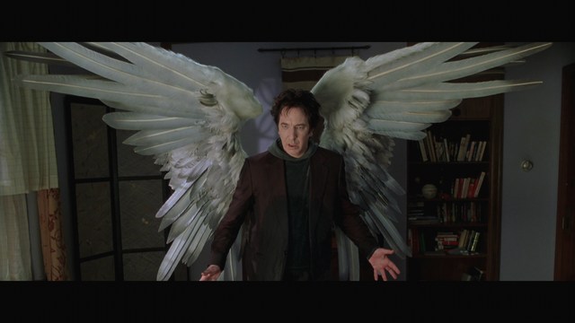 A man with large white wings stands in a room, wearing a dark suit, with a serious expression on his face. Bookshelves and curtains are visible in the background.