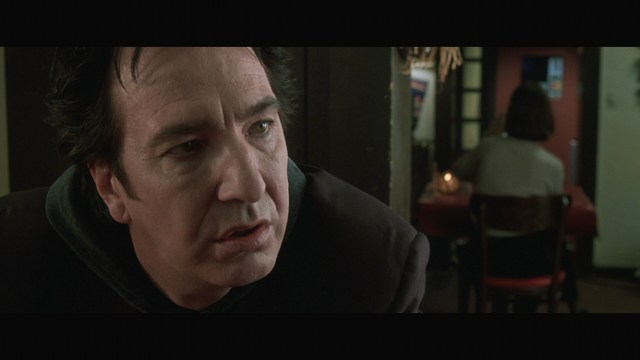 Close-up of a man with a serious expression, seated in a dimly lit setting, with another person in the background appearing to be seated at a table.