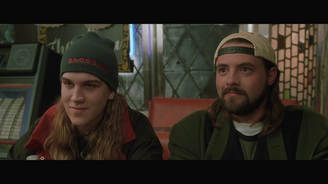 Two men sitting close together in a diner, one wearing a black beanie with the word 'SNOOGANS' and the other wearing a cap, both appearing engaged in conversation.
